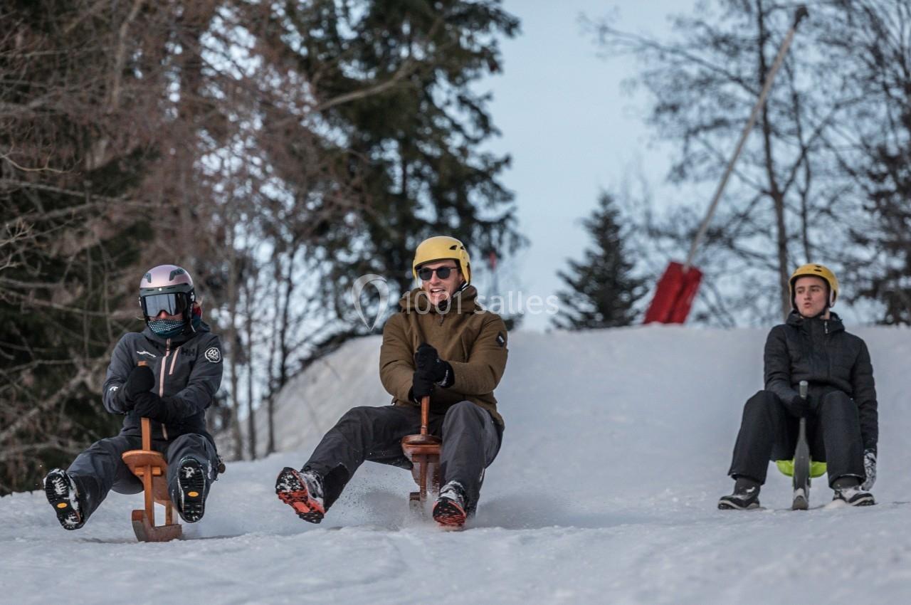 Trois personnes descendent une pente enneigée sur des luges en bois, entourées d'arbres en arrière-plan.