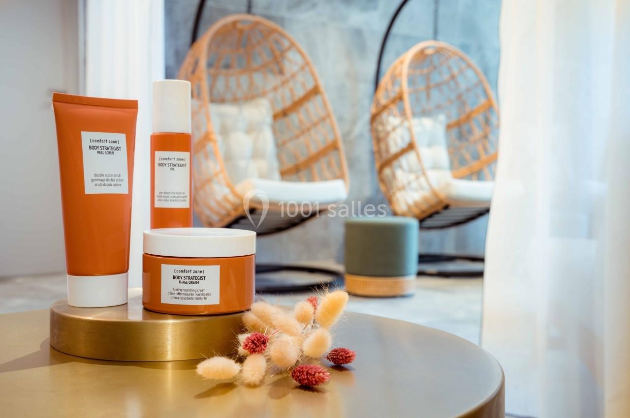 Produits cosmétiques sur une table dorée avec des fauteuils suspendus et des fleurs séchées en arrière-plan.