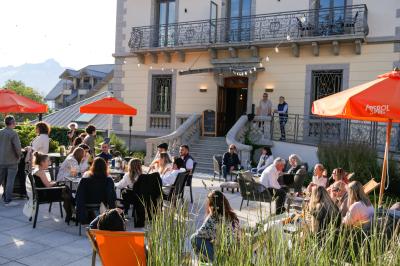 Façade d'un bâtiment éclairé la nuit, entouré d'un jardin avec des arbres, des chaises et des tables en extérieur.
