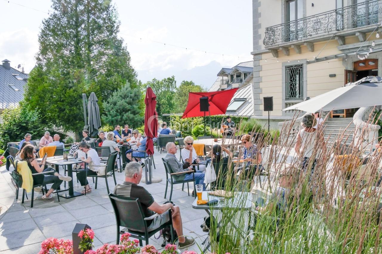 Terrasse extérieure d'un café avec des clients assis à des tables, entourée de végétation et de bâtiments.