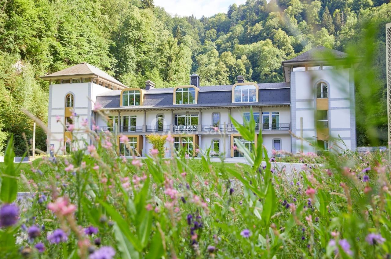 Bâtiment entouré de verdure avec des fleurs sauvages au premier plan et une forêt en arrière-plan.