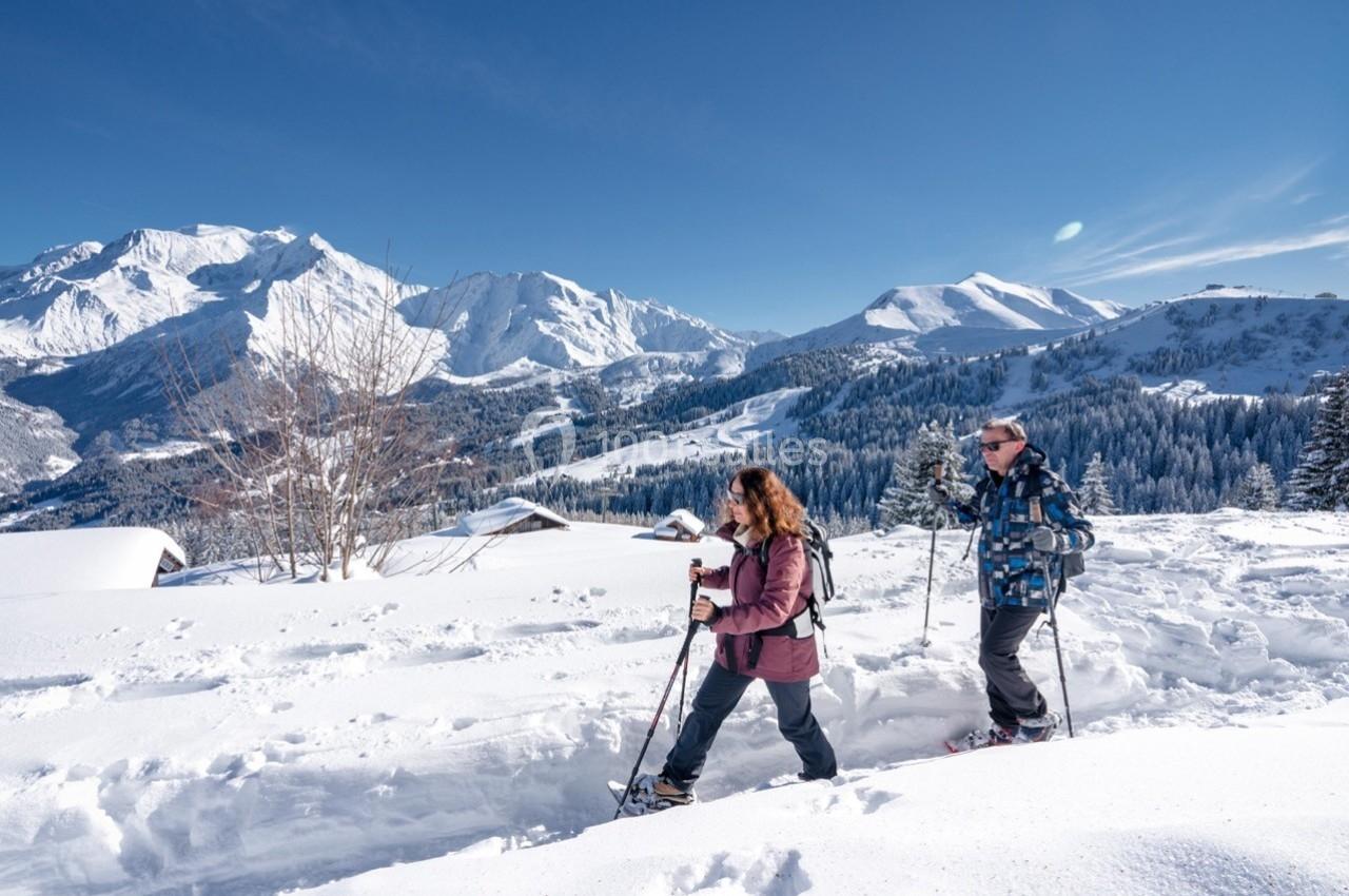 Deux personnes marchant en raquettes sur un sentier enneigé, avec des montagnes et chalets en arrière-plan.