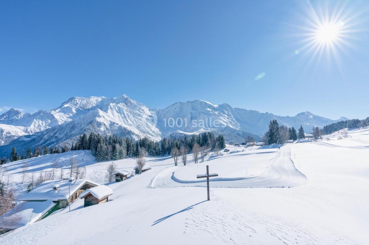 Paysage enneigé avec montagnes en arrière-plan, chalets dispersés et ciel dégagé sous un soleil éclatant.