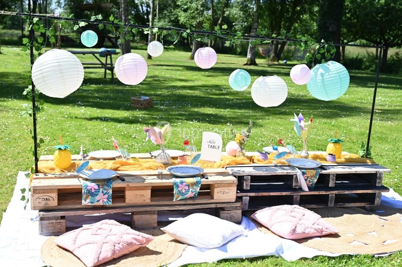 Table basse en palettes décorée pour un pique-nique, avec lanternes suspendues, coussins et vaisselle colorée dans un jardin.