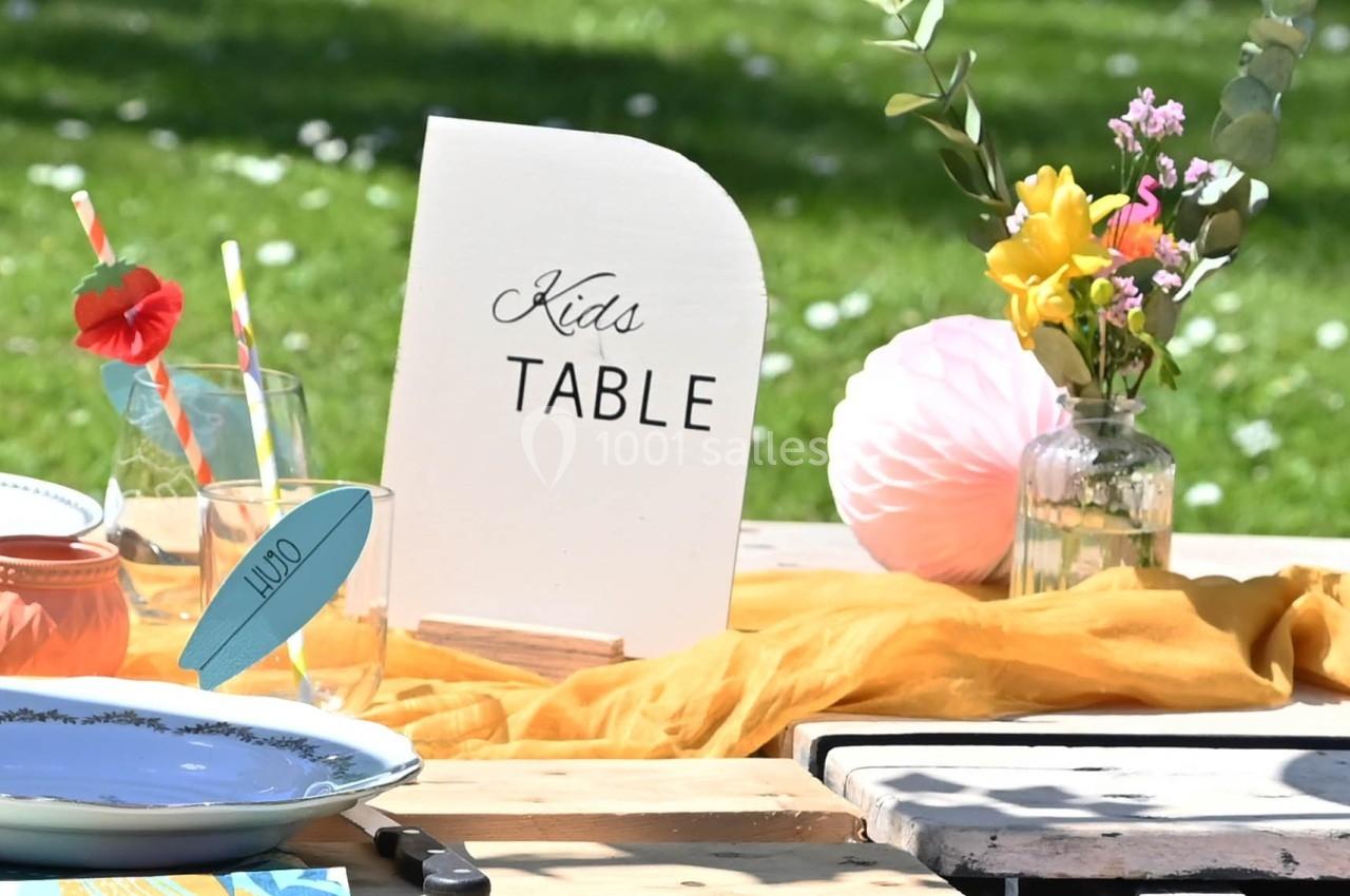 Table décorée pour enfants avec vaisselle colorée, fleurs, pancarte ’Kids Table’ et nappe jaune, en extérieur.