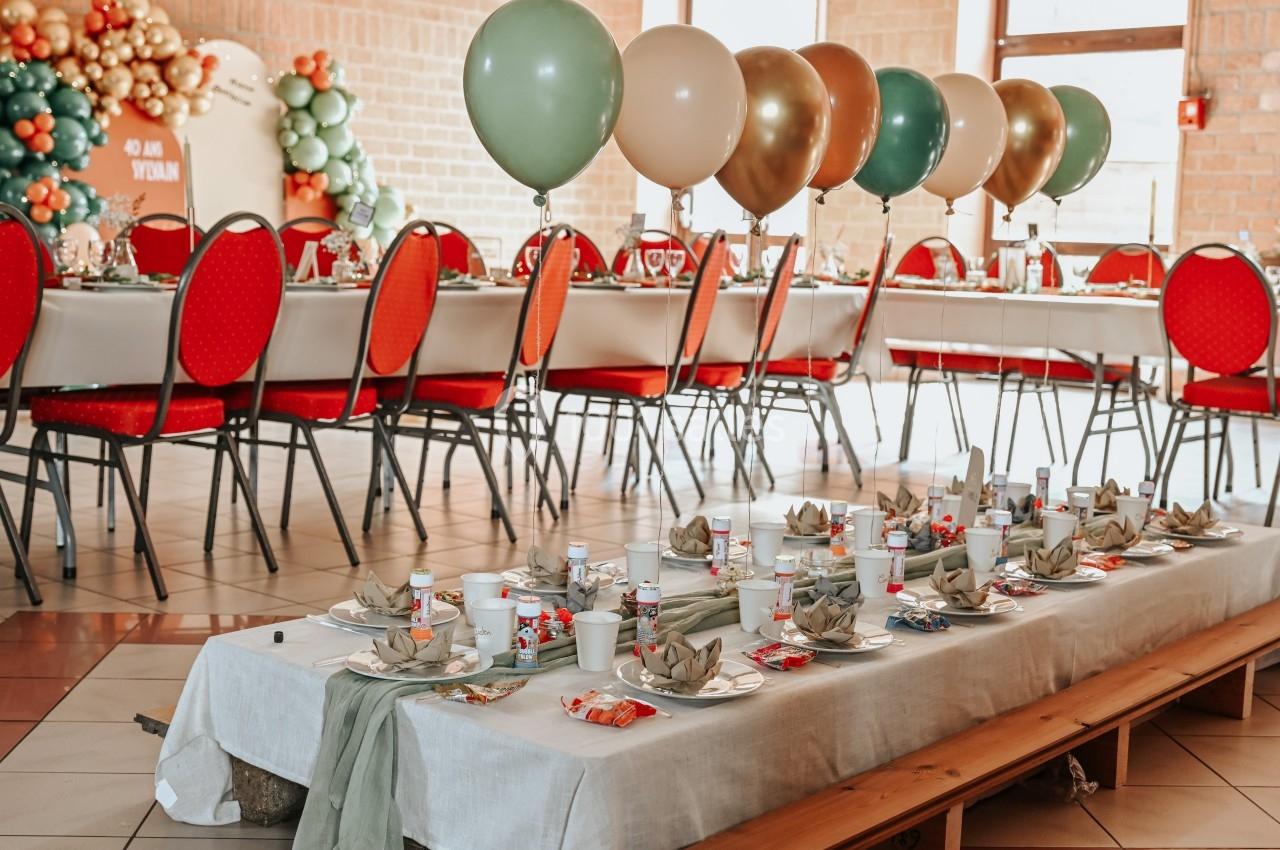 Table décorée pour un événement avec vaisselle, serviettes pliées et ballons colorés en arrière-plan.