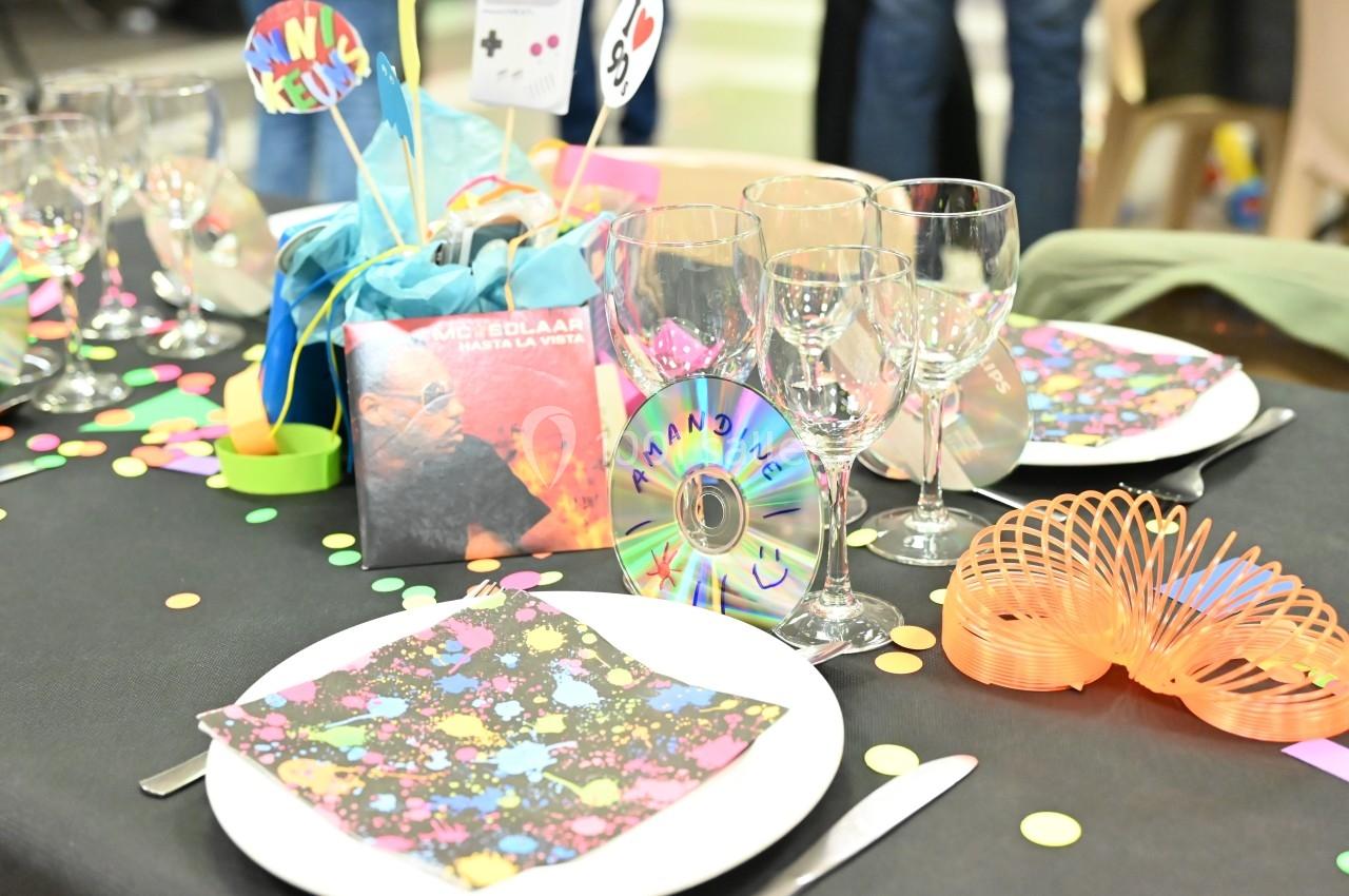 Table décorée avec des assiettes, serviettes colorées, verres, confettis, un CD et des éléments festifs.