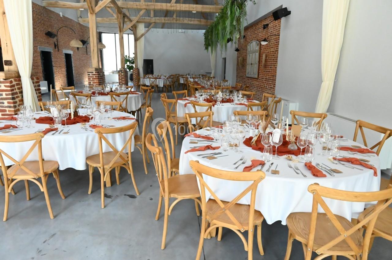 Salle de réception lumineuse avec tables rondes dressées, nappes blanches, serviettes rouges et chaises en bois clair.