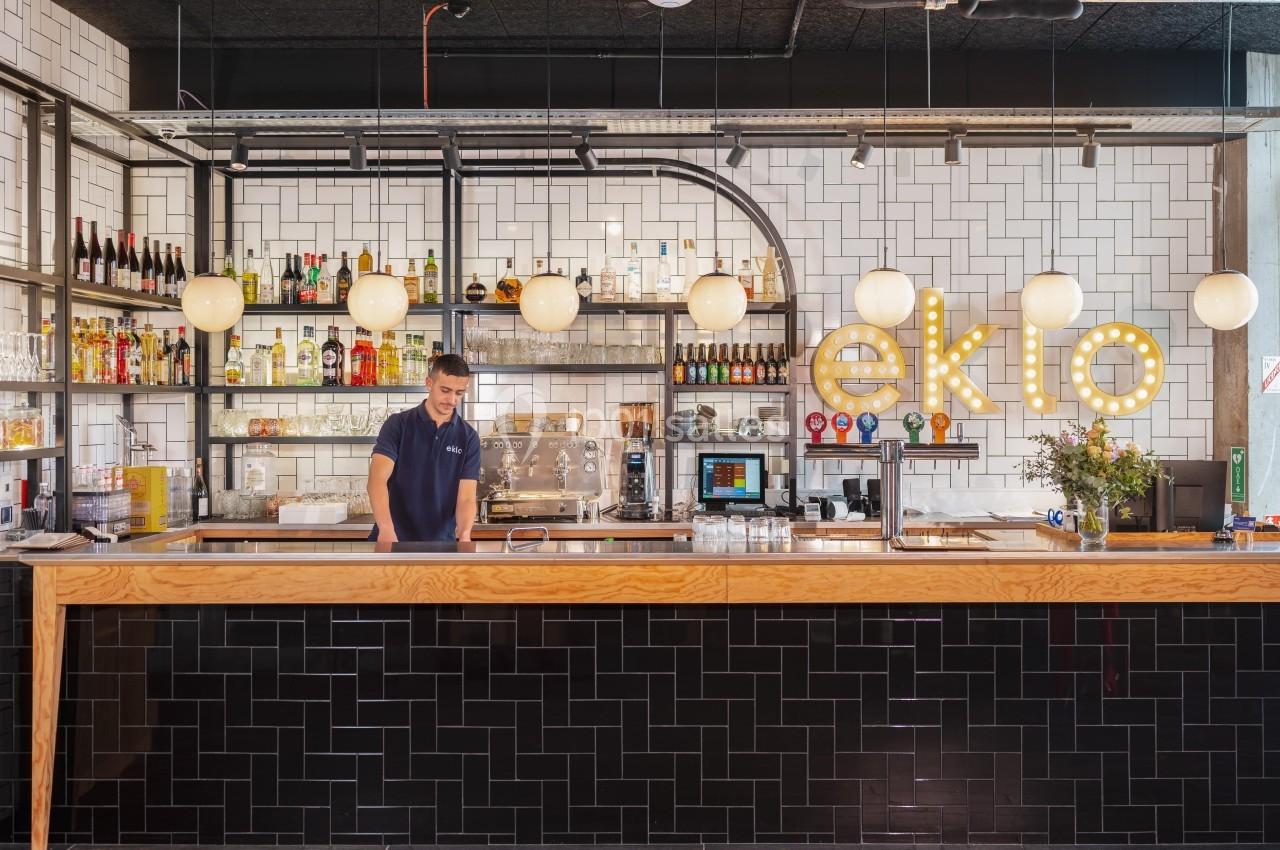 Bar moderne avec comptoir en bois, carrelage mural blanc, étagères garnies de bouteilles et lumières suspendues.