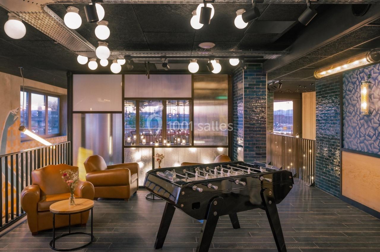 Salle de détente moderne avec baby-foot, fauteuils en cuir, éclairage suspendu et décoration contemporaine.