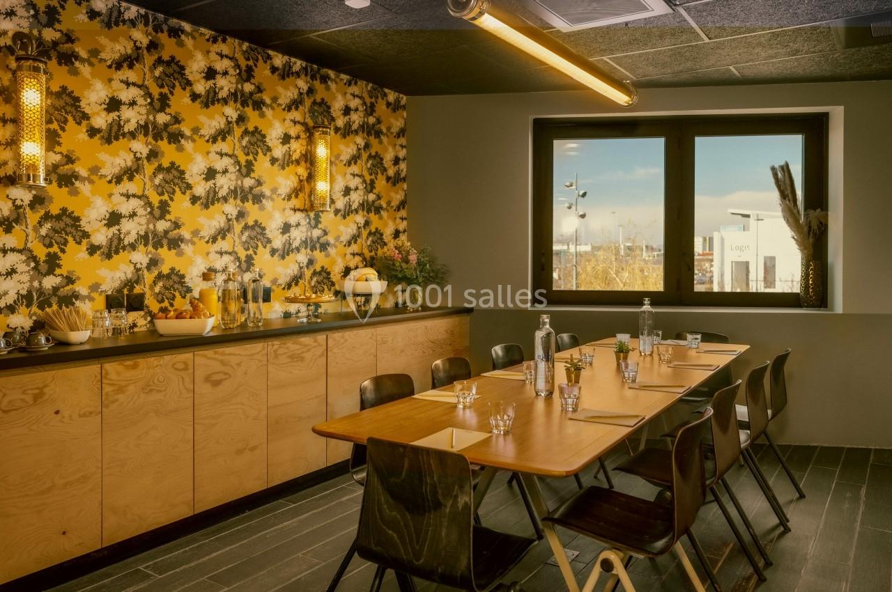 Salle de réunion lumineuse avec table en bois, chaises noires, buffet, mur à motif floral et grande fenêtre.
