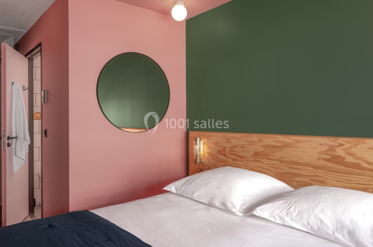 Chambre avec un mur rose et vert, un miroir rond, un lit double avec draps blancs et une salle de bain attenante.