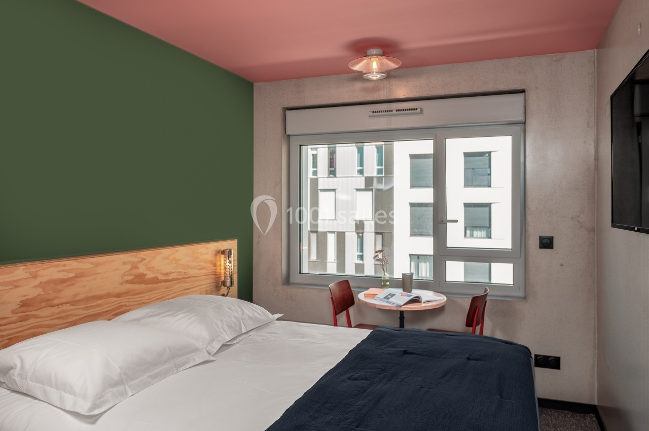 Chambre d'hôtel moderne avec lit double, mur vert, table ronde près d'une fenêtre donnant sur des bâtiments.