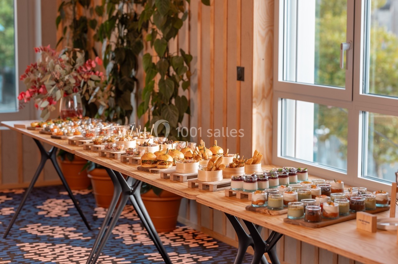 Table en bois garnie de plats variés, mini-burgers, verrines et amuse-bouches, dans une salle lumineuse avec plantes.