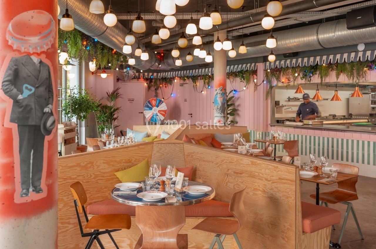 Salle de restaurant moderne avec tables en bois, décoration colorée, luminaires suspendus et cuisine ouverte en arrière-plan.