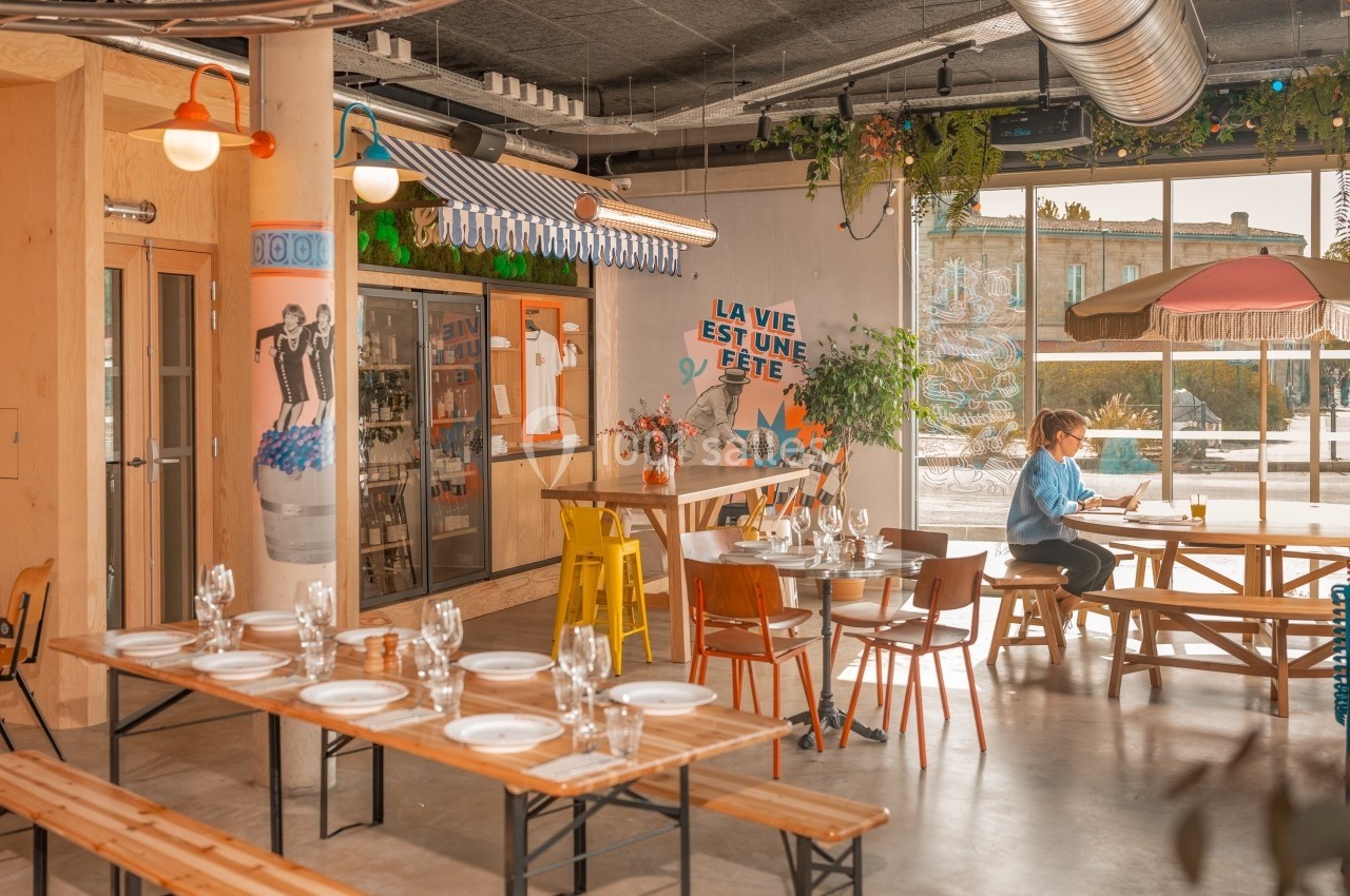 Salle de restaurant lumineuse avec tables en bois, vaisselle dressée, décor coloré et une personne assise près d'une fenêtre.