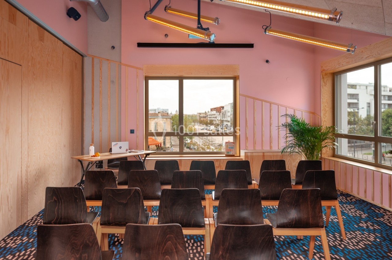 Salle de réunion lumineuse avec des chaises en bois, une table, des murs roses et une vue sur des bâtiments urbains.