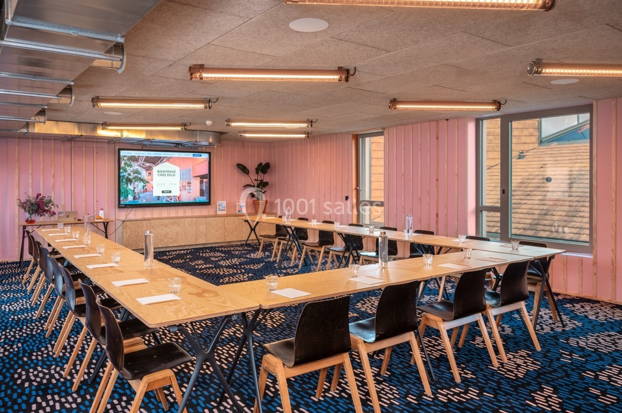 Salle de réunion lumineuse avec grandes tables en bois disposées en U, chaises noires et écran de présentation.