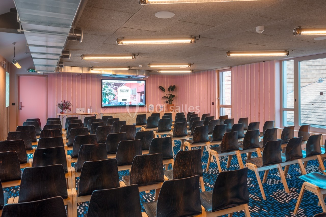 Salle de conférence moderne avec rangées de chaises en bois, écran allumé et murs en bois clair.