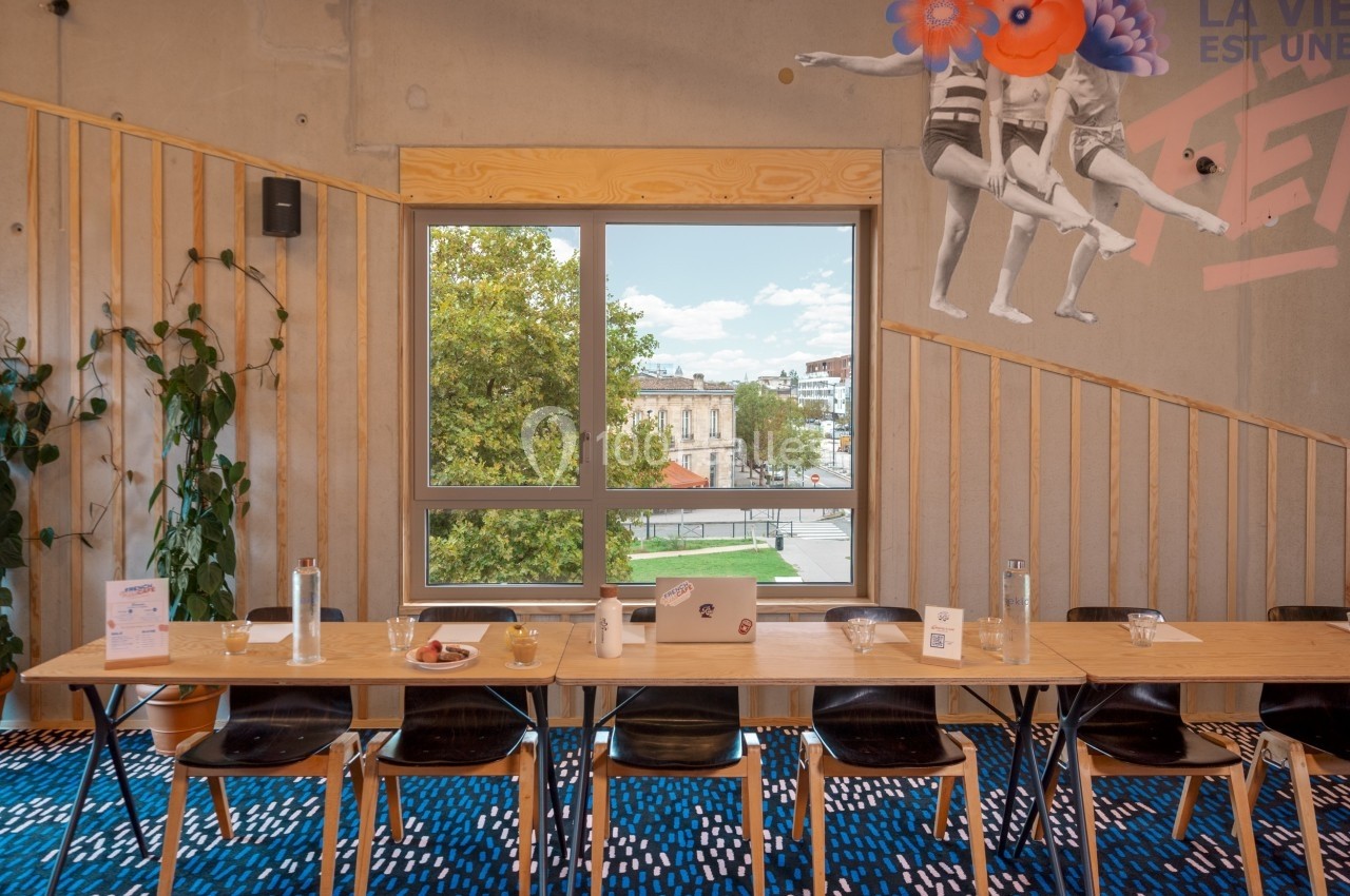 Salle de réunion lumineuse avec une grande table, des chaises, une fenêtre donnant sur un paysage urbain et une fresque…