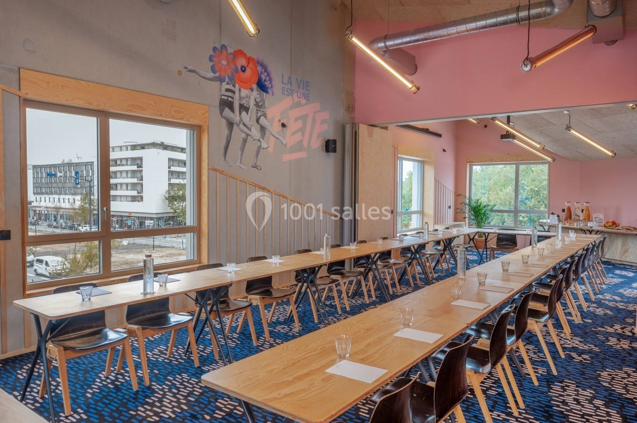 Salle de réunion lumineuse avec de longues tables en bois, chaises noires et vue sur l'extérieur par de grandes fenêtres.