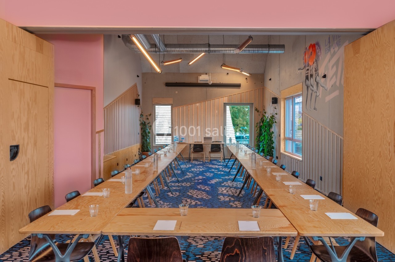 Salle de réunion lumineuse avec tables disposées en U, chaises en bois, murs décorés et grandes fenêtres.