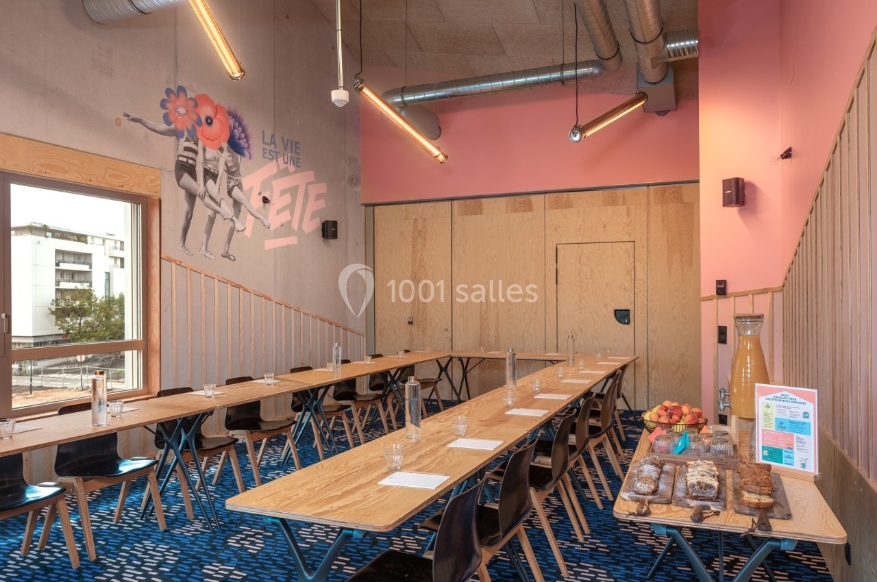 Salle de réunion lumineuse avec tables en bois, chaises noires, mur rose et buffet de collations sur le côté.