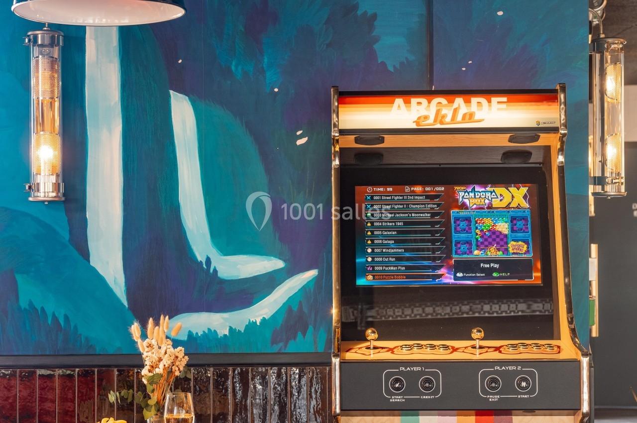 Borne d'arcade rétro près d'une table avec des boissons et des snacks, dans un décor coloré avec une fresque murale.