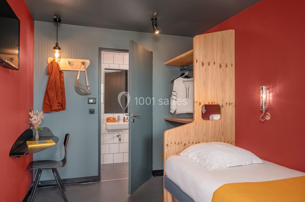 Chambre moderne avec lit simple, bureau, étagère en bois, et vue sur une salle de bain carrelée avec miroir.