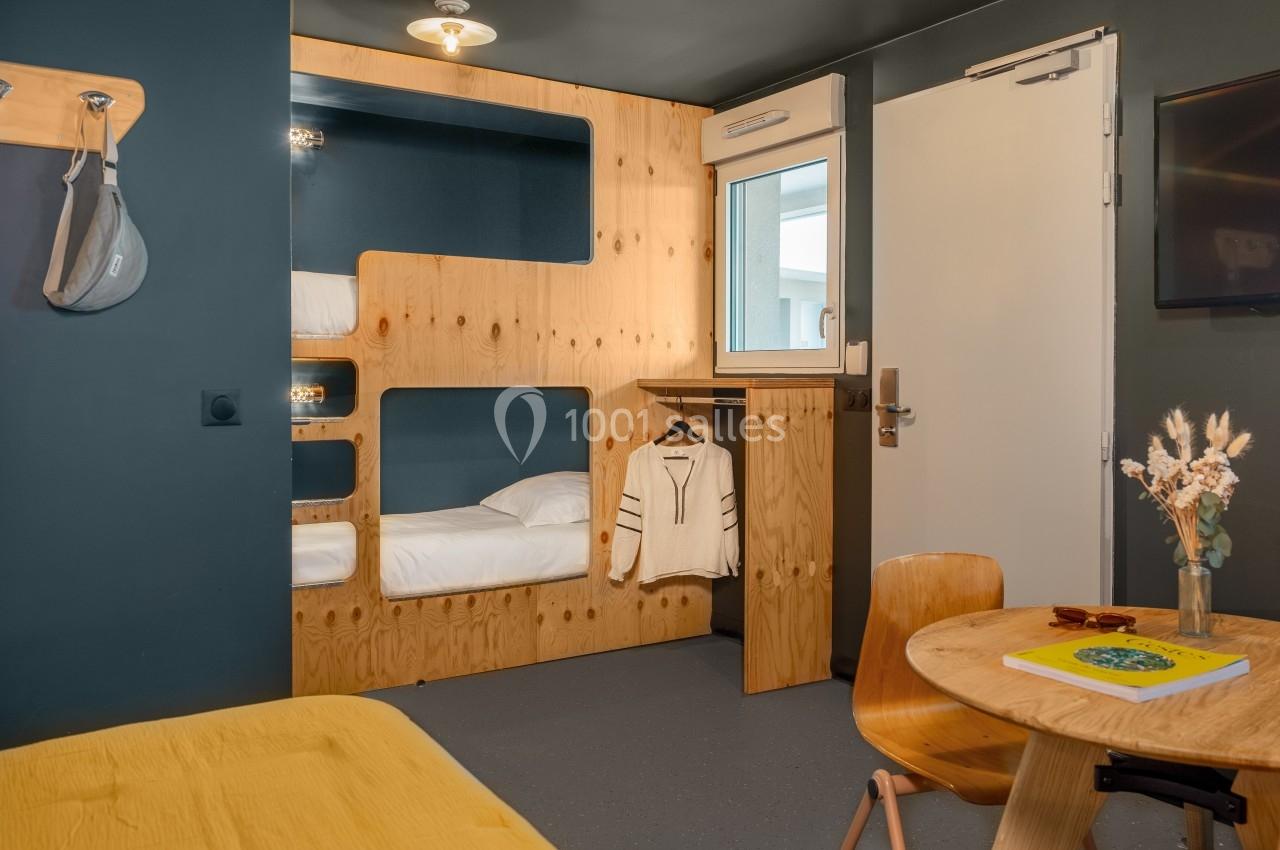 Chambre moderne avec lits superposés en bois, table ronde, chaise, et porte donnant sur l'extérieur.