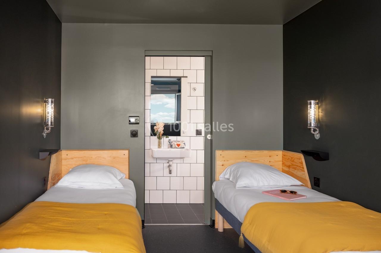Chambre avec deux lits simples, draps jaunes, murs sombres et vue sur une salle de bain carrelée avec lavabo.