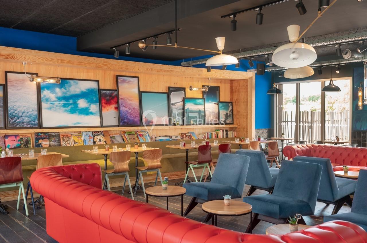 Espace lounge coloré avec canapés rouges, fauteuils bleus, tables basses et murs décorés de cadres et étagères.