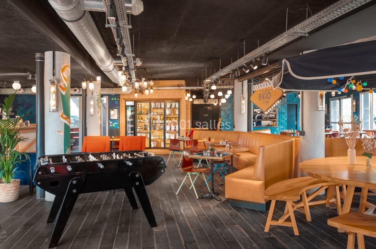 Salle conviviale avec baby-foot, banquettes en cuir, tables en bois et éclairage moderne dans un espace lumineux.