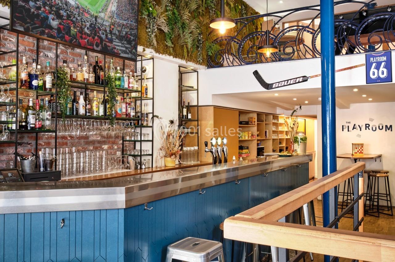 Bar moderne avec comptoir bleu, étagères de bouteilles, écran diffusant un match et décoration végétale au mur.