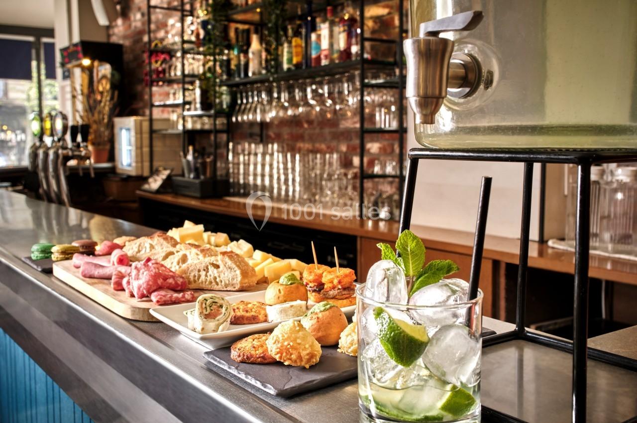 Plateau de charcuterie, fromages et amuse-bouches sur un comptoir de bar avec un verre de cocktail en premier plan.