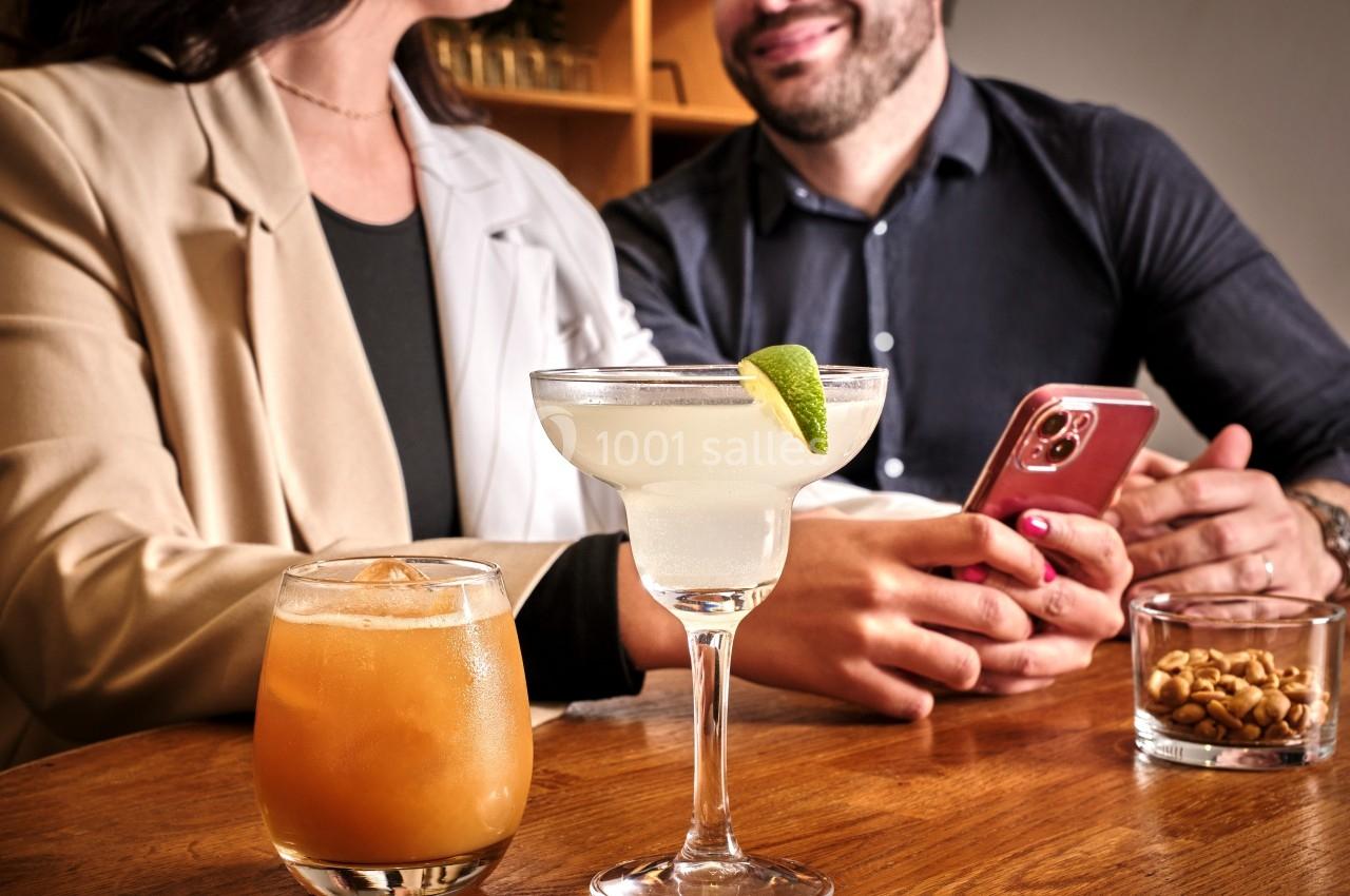 Deux personnes assises à une table avec des cocktails et des cacahuètes, l'une tenant un téléphone.