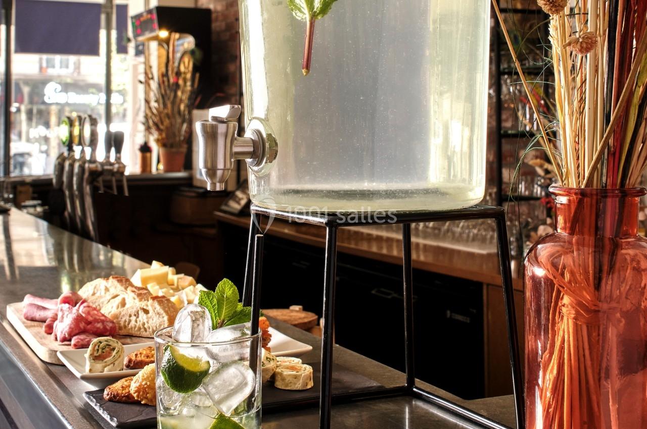 Distributeur de boisson citronnée sur pied, avec plateau de charcuterie et verre garni de menthe au premier plan.