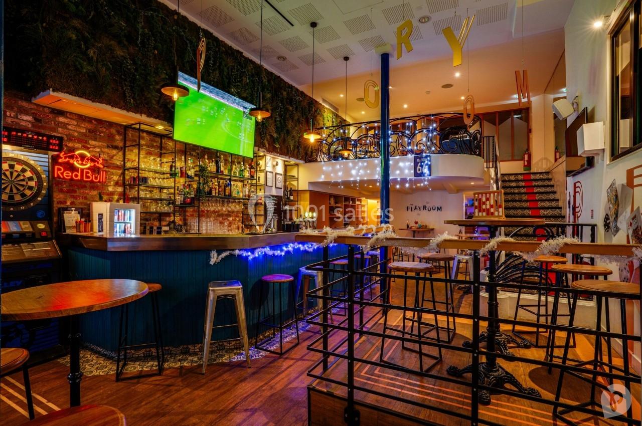 Intérieur d'un bar moderne avec comptoir éclairé, tables hautes, écran diffusant un match et décoration lumineuse.