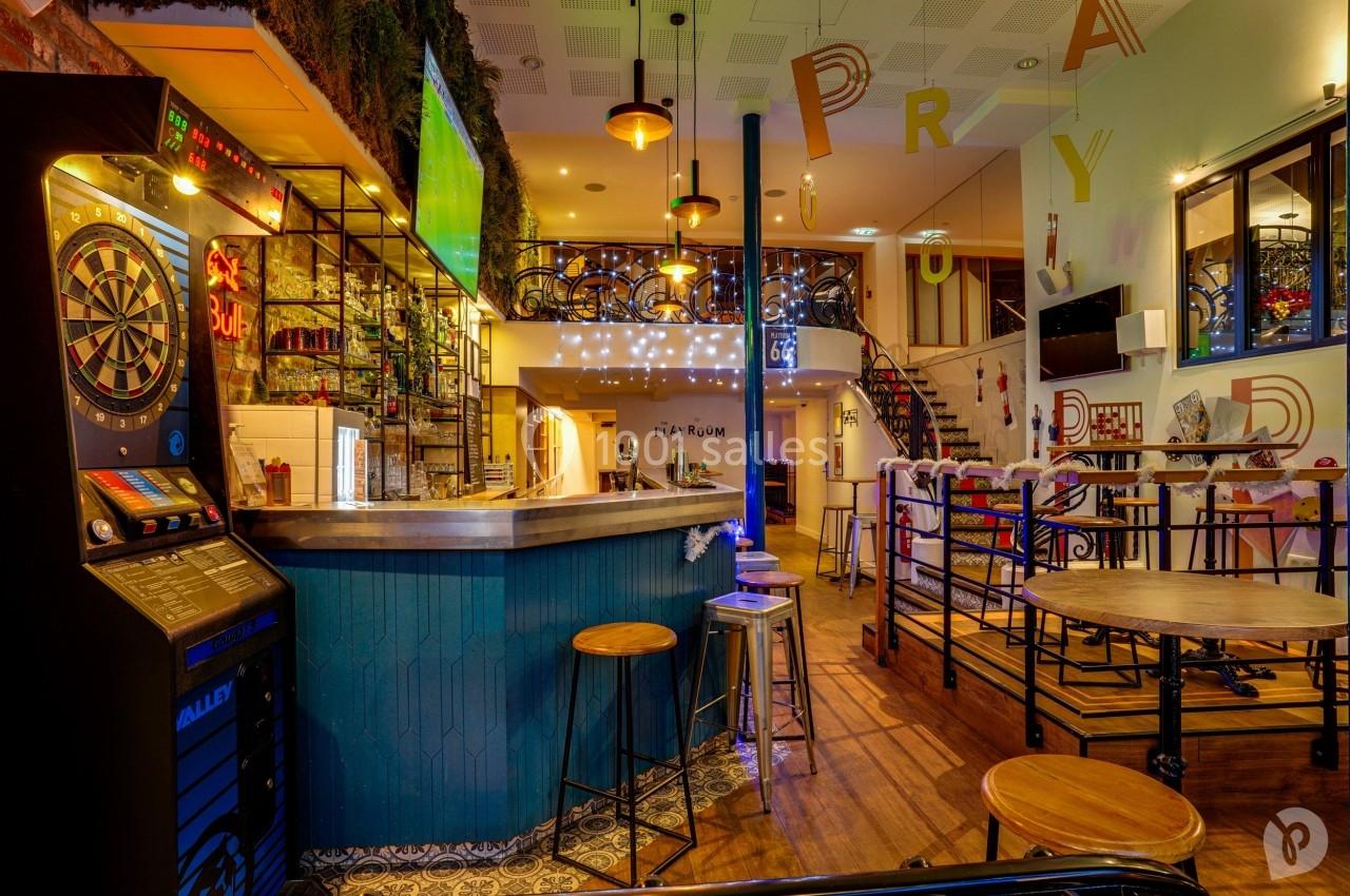 Bar avec comptoir en bois, jeux de fléchettes, tables hautes, escalier en colimaçon et décorations lumineuses.