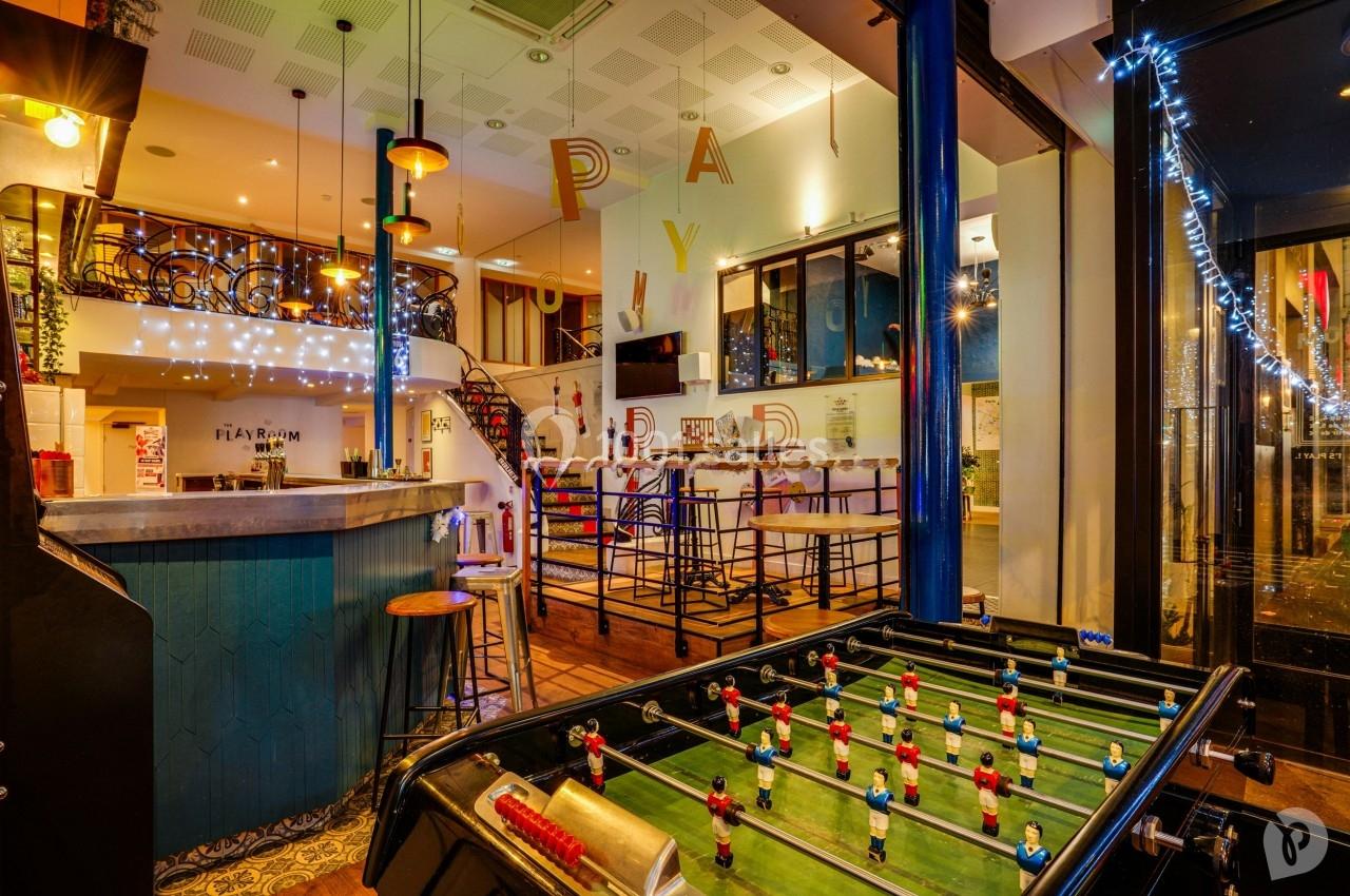 Bar lumineux avec baby-foot au premier plan, tables hautes et décoration colorée en arrière-plan.