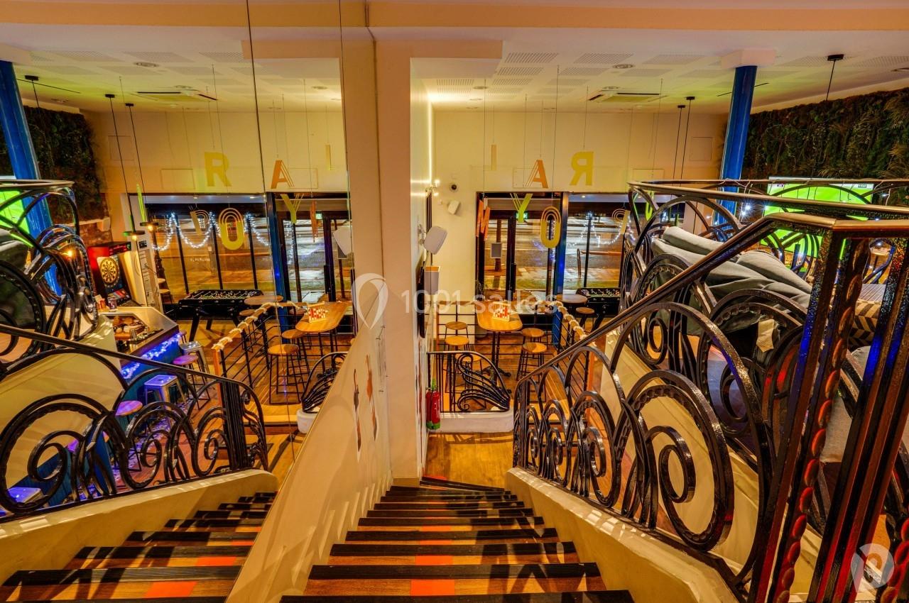 Escalier intérieur avec rambardes en fer forgé, menant à une salle lumineuse avec tables et grandes baies vitrées.