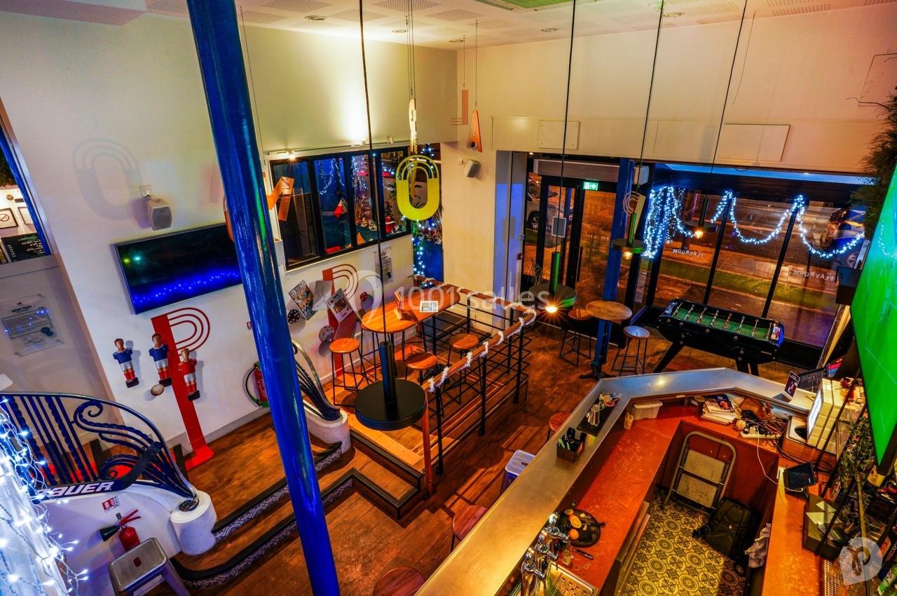 Vue intérieure d'un bar coloré avec un comptoir, des tables hautes, un baby-foot et des décorations lumineuses.