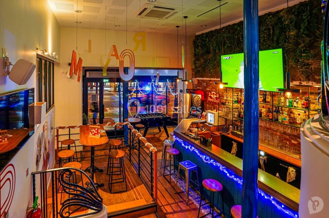 Intérieur d'un bar avec comptoir éclairé, tables hautes, baby-foot, cible de fléchettes et écrans diffusant un match.