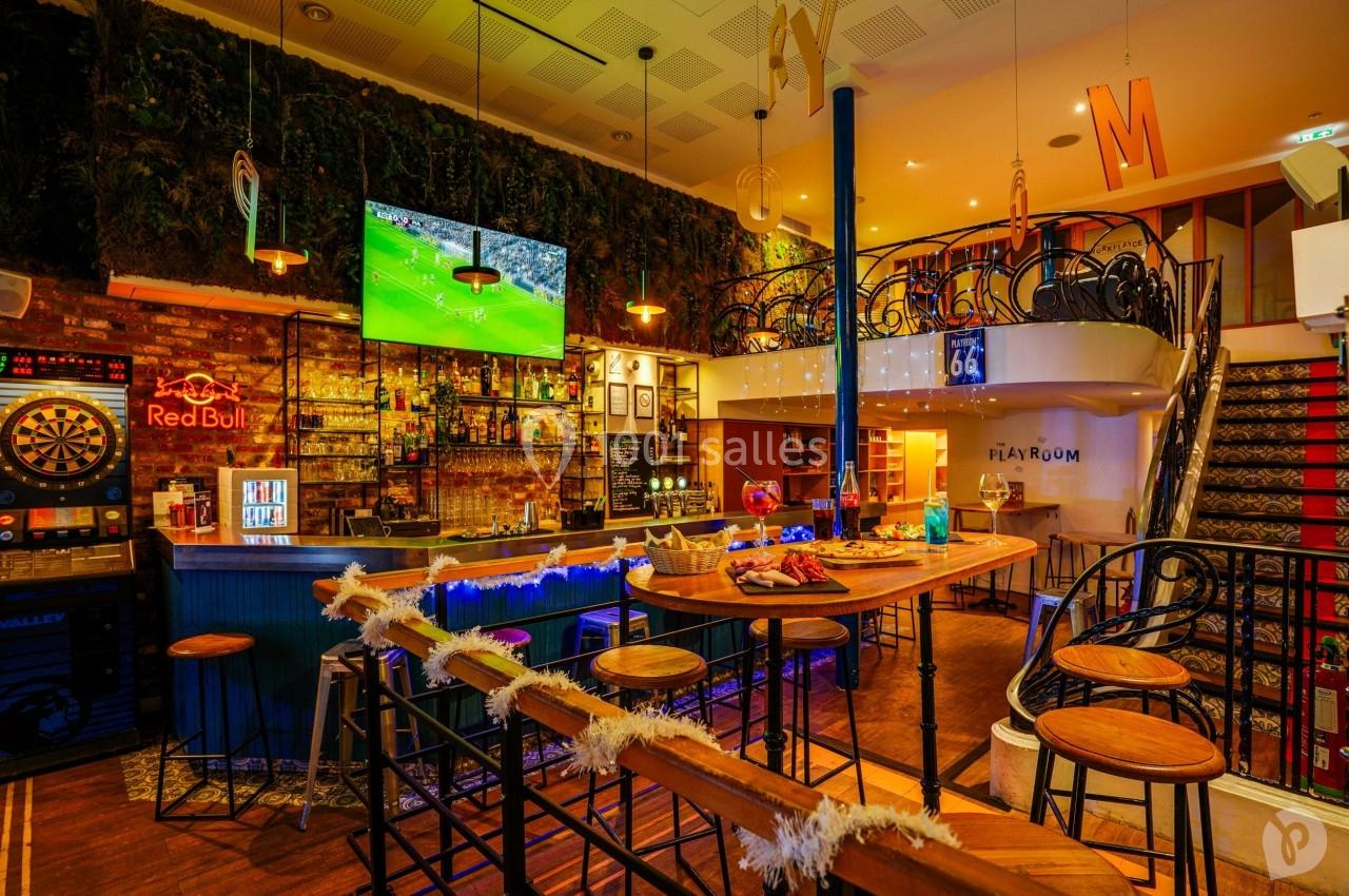 Intérieur d'un bar avec comptoir éclairé, tables hautes, écran diffusant un match et décoration chaleureuse.