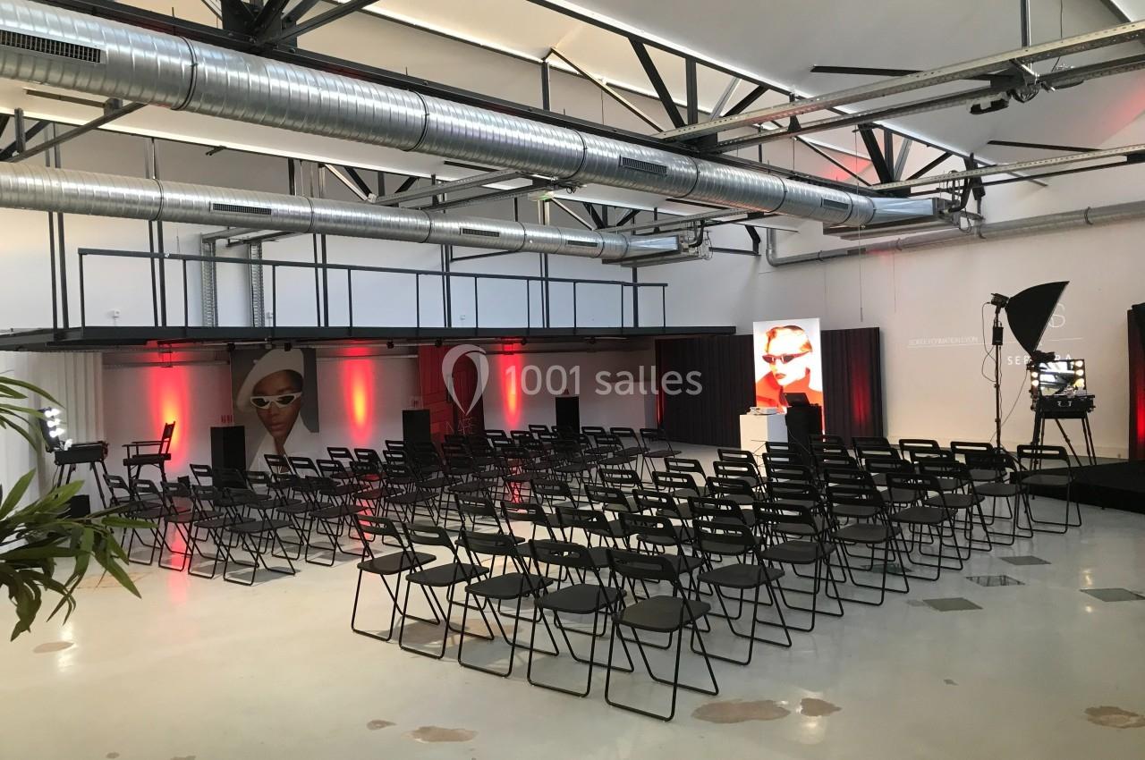 Salle aménagée pour un événement avec des rangées de chaises noires, éclairage rouge et affiches publicitaires.