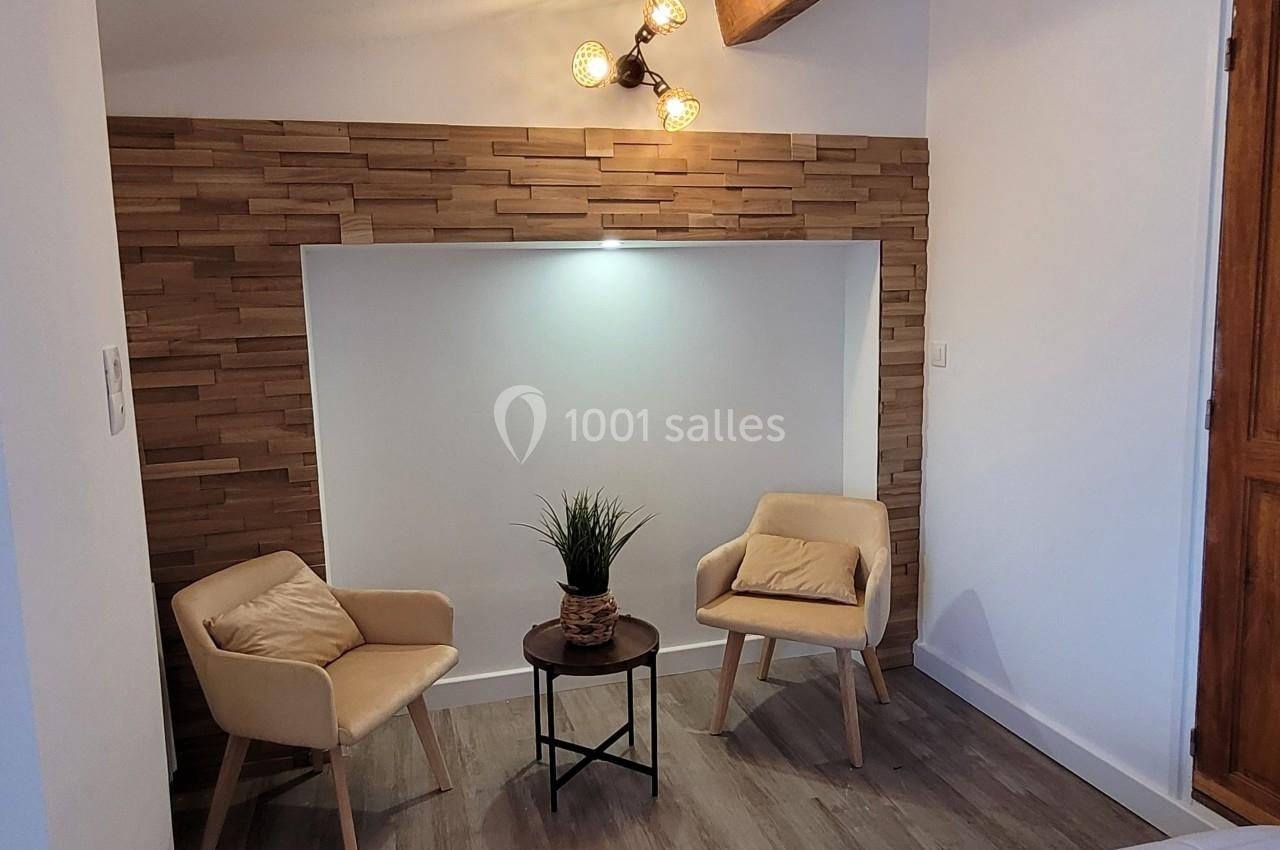 Deux fauteuils beige autour d'une table basse avec une plante, dans un coin décoré de bois et éclairé par une applique…