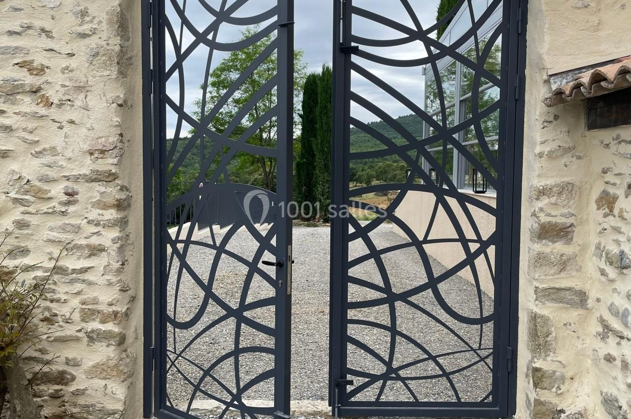 Portail métallique noir ajouré avec motifs abstraits, encadré par des murs en pierre, donnant sur une allée gravillonnée.