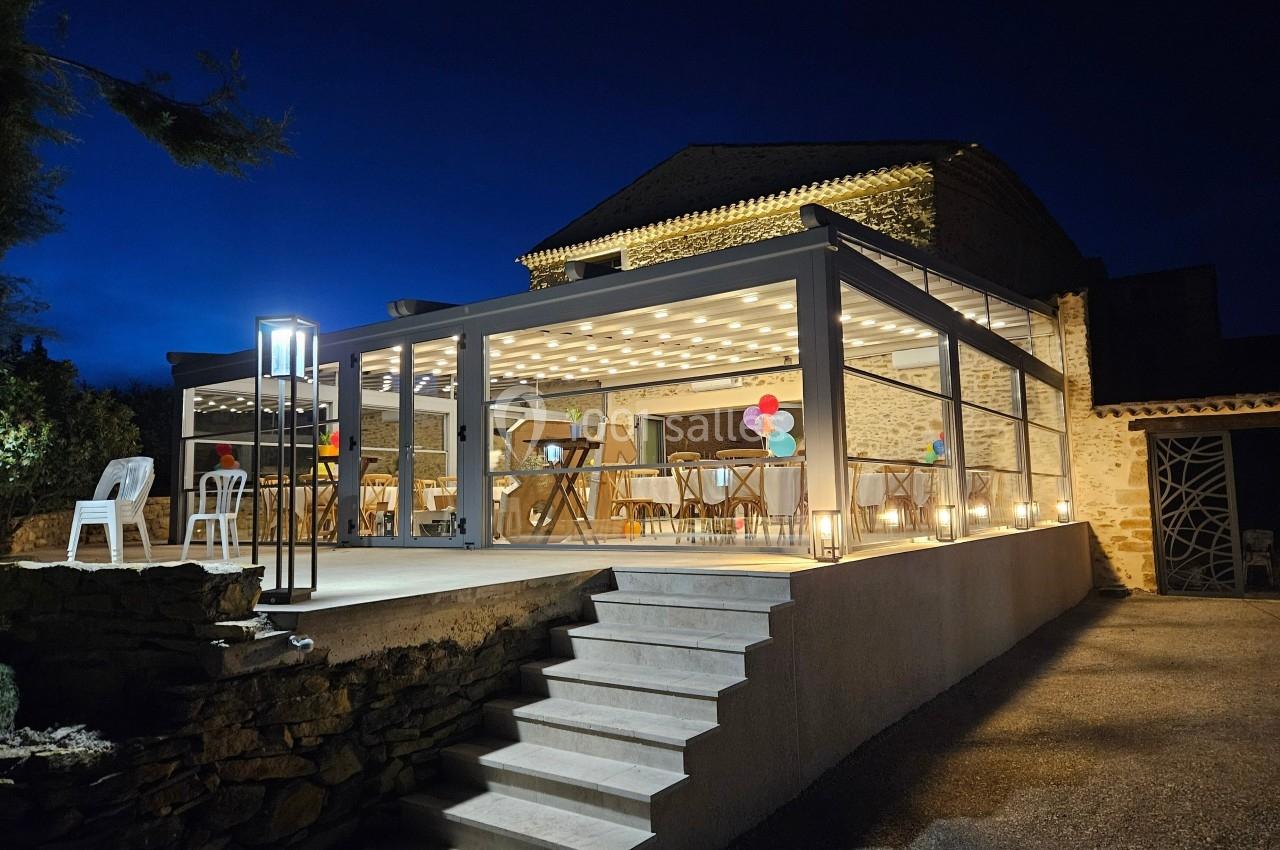 Terrasse éclairée avec une pergola vitrée, décorée de ballons colorés, devant un bâtiment en pierre, de nuit.