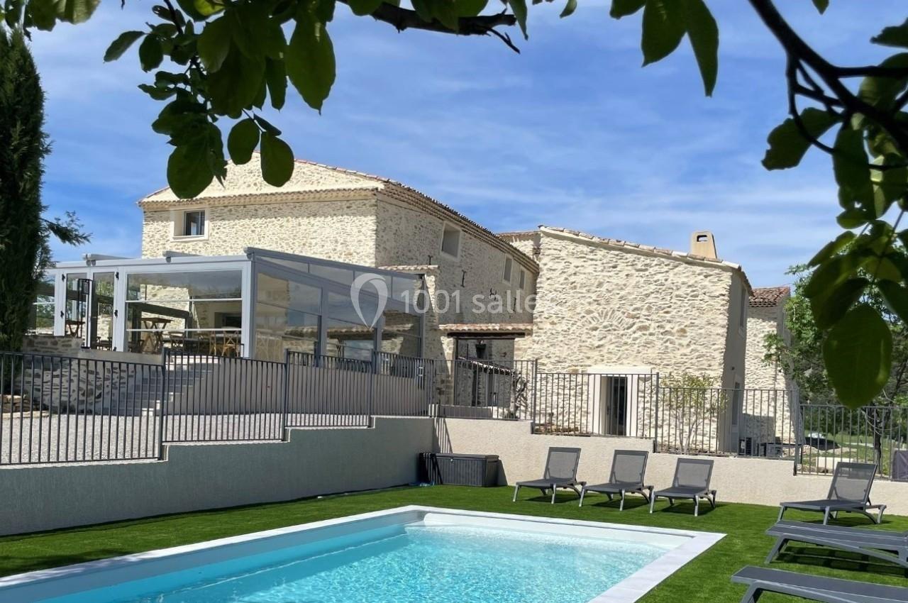 Maison en pierre avec piscine, terrasse vitrée et chaises longues, entourée de végétation sous un ciel dégagé.