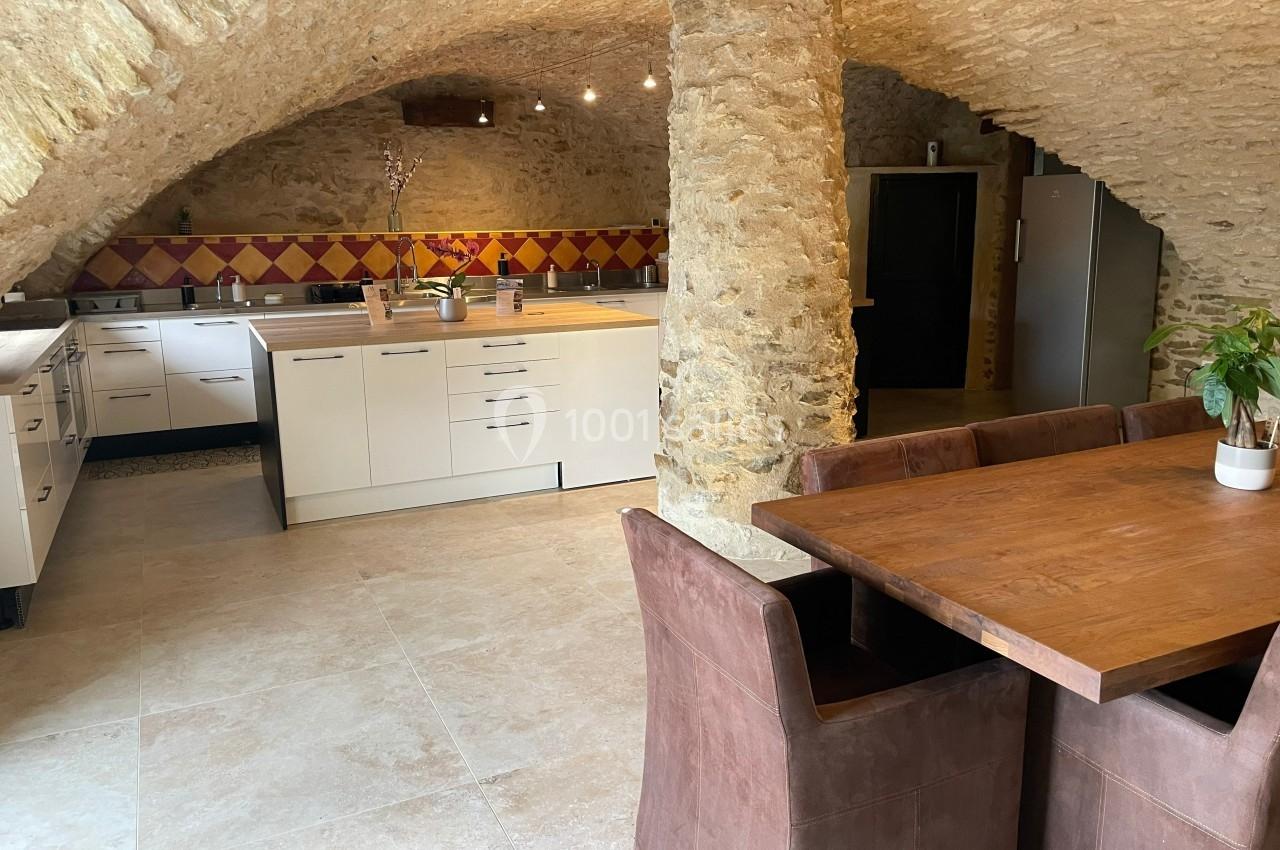 Cuisine aménagée sous une voûte en pierre, avec meubles modernes, table en bois et chaises en tissu marron.